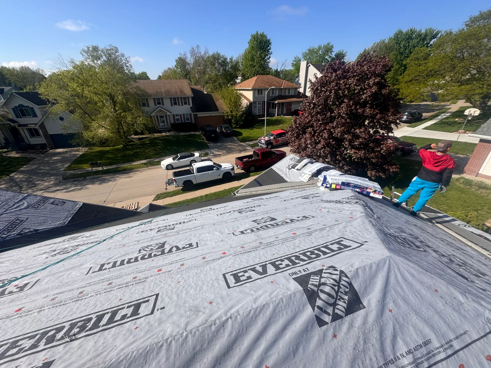 Crew installing white Everbilt synthetic roof underlayment on sloped roof deck with Timberline HD shingle bundles staged at ridge
