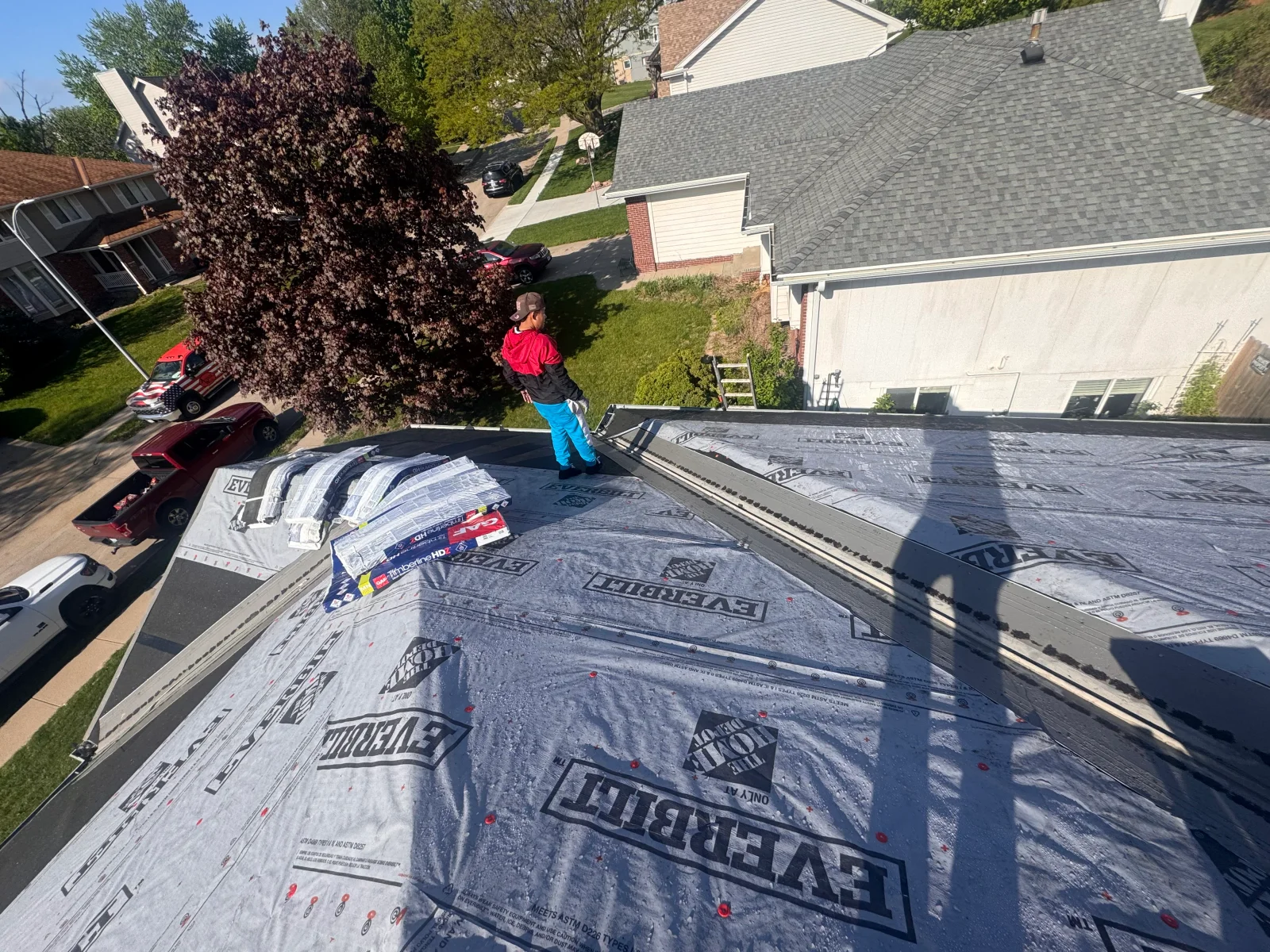 Worker on steep roof installing Everbilt synthetic underlayment with red cap nails and stacked underlayment rolls